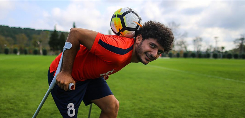 Resim: Kırmızı formasıyla sahada duran amputee bir futbolcu, koltuk değnekleriyle dengede dururken topu ensesinde ustalıkla tutuyor. Arkada yeşil bir saha ve ağaçlar görülüyor.