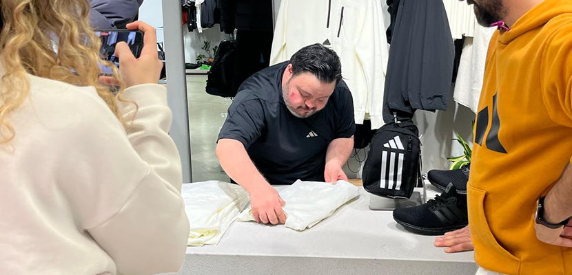 Betimleme: Fotoğrafta, down sendromlu Berk Canbulat bir mağazada çalışan bir kişi tezgâh üzerinde kıyafet katlıyor. Üzerinde siyah bir tişört var ve dikkatle işine odaklanmış. Sağda turuncu kapüşonlu bir kişi onu izliyor, solda ise bir kişi telefonuyla bu anı görüntülüyor. Arka planda askılarda spor kıyafetler ve bir sırt çantası yer alıyor. Ortam modern ve düzenli görünüyor.