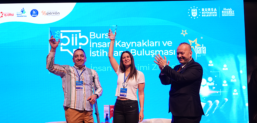 Sahnedeki karede, solda Mehmet Kızıltaş, elindeki cam ödülü gururla havaya kaldırırken güçlü ve kendinden emin bir duruş sergiliyor. Ortada Güler Koca, aynı şekilde ödülünü yukarı kaldırmış, tebessümüyle sahnenin coşkusunu paylaşan bir görünümde. Sağda ise zirve yetkilisi, iki ödül sahibine dönük şekilde alkışlayarak onları tebrik ediyor ve sahnenin resmi, destekleyici atmosferini tamamlıyor. Arka planda “Bursa İnsan Kaynakları ve İstihdam Buluşması” temalı mavi fon, etkinliğin kurumsal logosu ve organizasyonun profesyonel yapısını yansıtan görseller yer alıyor. Genel olarak kare, başarı, takdir ve emekle kazanılmış bir gurur anını güçlü biçimde yansıtıyor.
