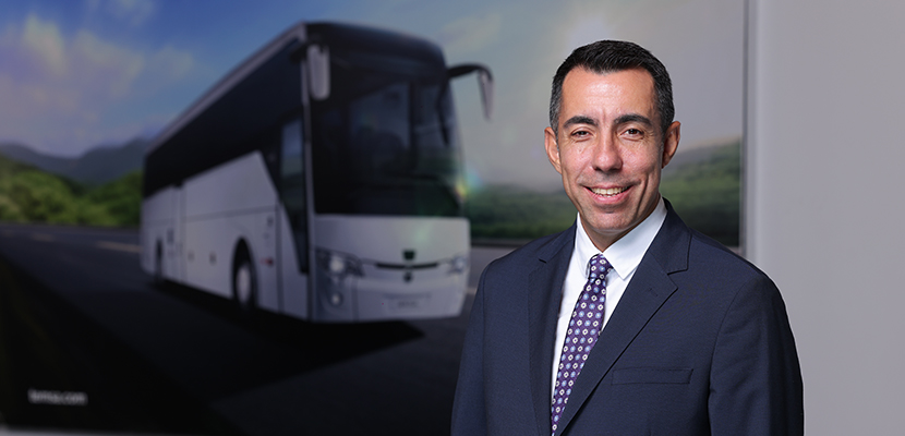 Resim Betimleme: Erhan Özel, koyu renk takım elbise ve kravat giymiş. Ayakta duruyor. Arka planda büyük beyaz bir otobüs var. Otobüsün çevresinde yeşil ağaçlar ve dağlar görünüyor. Hava açık ve gündüz. Zemin asfalt.