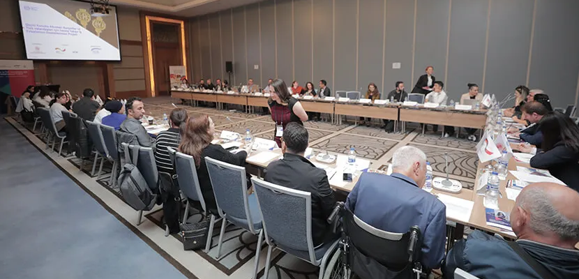 Betimleme: Otel toplantı salonunda U şeklindeki masa etrafından katılımcılar yan yana otuyor. Sahnede açık parkovizyon sunumu var.