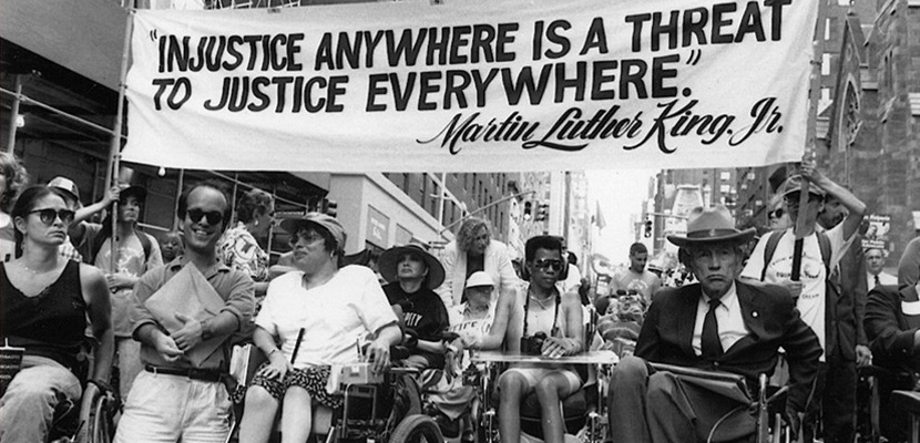 Betimleme: Bir grup engelli aktivist, tekerlekli sandalyeleriyle şehir merkezinde bir protesto yürüyüşünde ilerliyor. Kalabalığın önünde büyük bir pankart taşınıyor. Pankartta “Injustice anywhere is a threat to justice everywhere – Martin Luther King Jr.” yazıyor. Bu sözün Türkçesi: “Herhangi bir yerdeki adaletsizlik, her yerdeki adalet için tehdittir.” Yüksek binaların arasında ilerleyen grup, güçlü bir dayanışma, eşitlik ve hak savunuculuğu mesajı veriyor.