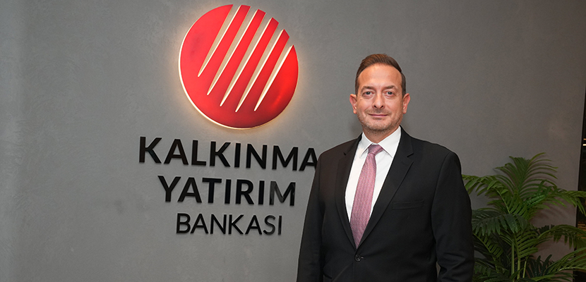 Betimleme: Kalkınma Yatırım Bankası Genel Müdür Yardımcısı Yasin Külahçı, kurum logosunun yer aldığı duvarın önünde profesyonel bir duruşla poz vermektedir. Takım elbisesi ve kurumsal ortam, üst düzey yöneticilik rolünü yansıtan ciddi ve temsil niteliği yüksek bir görünüm sunar.